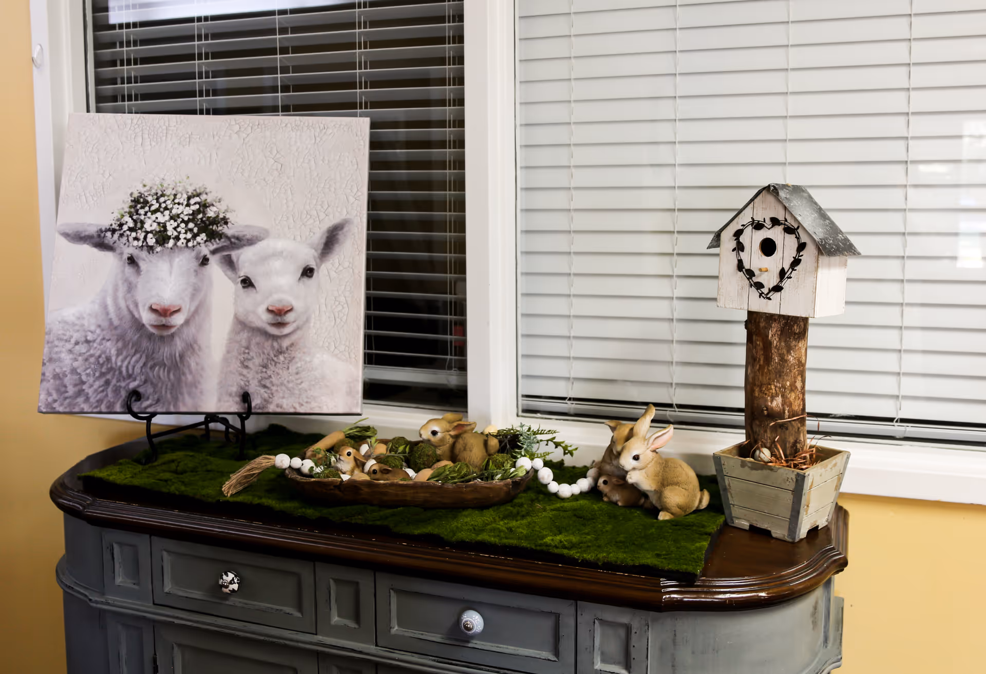 A decorative setup on a dark wooden sideboard with a green moss-like runner. On the left, there is a painting of two lambs, one wearing a flower crown. In the center, a wooden tray holds small rabbit figurines and white bead garland. On the right, a rustic birdhouse on a wooden post stands inside a square planter. The background features white window blinds.