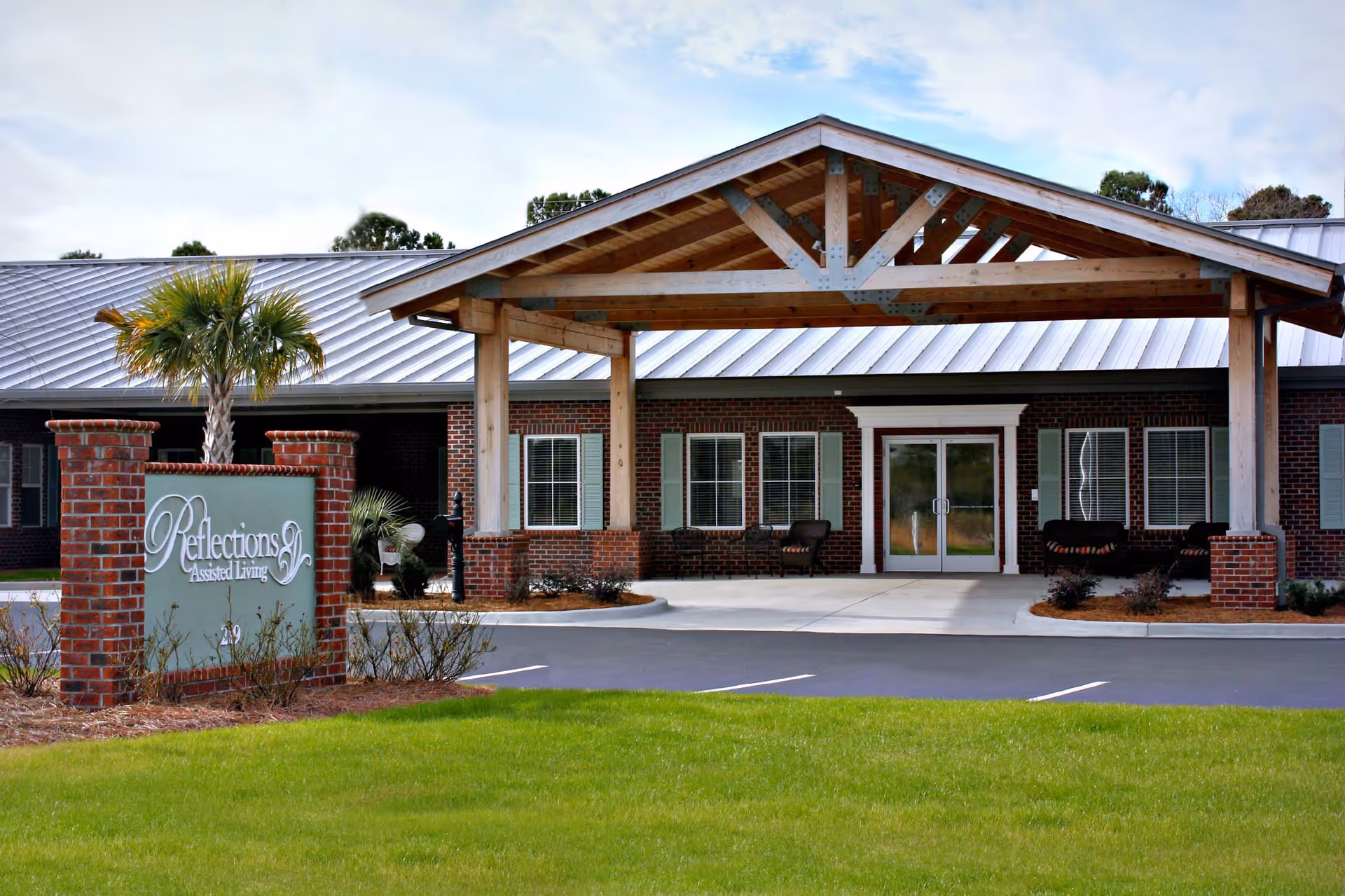 Exterior view of Reflections Assisted Living at Carolina Forest showing the entrance with a covered wooden canopy, brick pillars, and a sign with the facility's name. The building has a metal roof and several windows with shutters. There is a small palm tree and some landscaping in front.