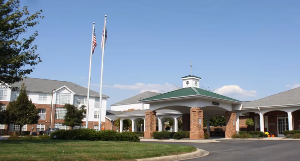 Front exterior of a senior living facility showing a covered entrance with brick columns, two flagpoles, and surrounding lawn.