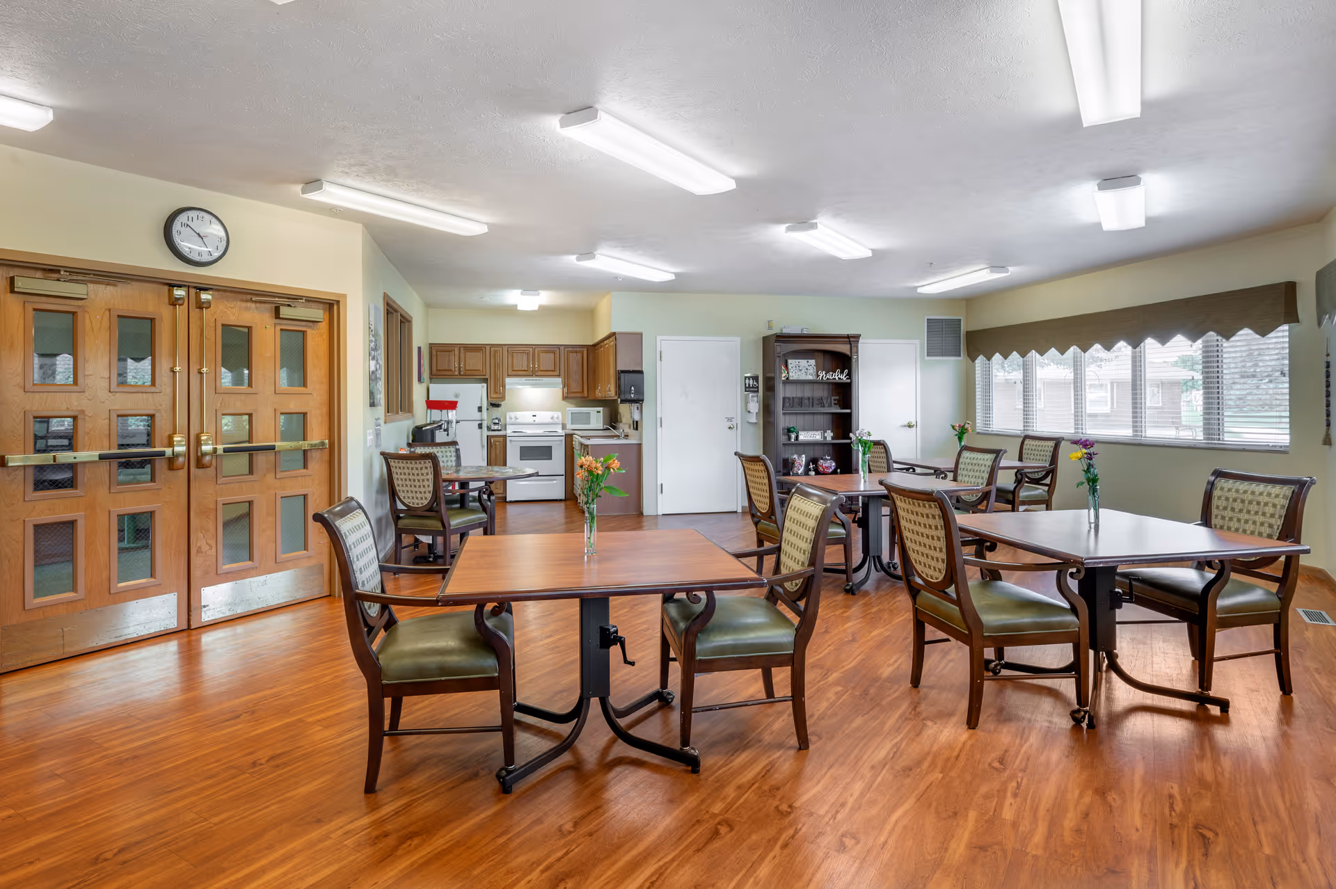 A well-lit dining area in a senior living facility with wooden floors, several square wooden tables each with two chairs, and small vases with flowers on the tables. In the background, there is a kitchen area with wooden cabinets, a stove, microwave, and refrigerator. Large windows with blinds allow natural light into the room. A clock is mounted above double wooden doors on the left side of the image.