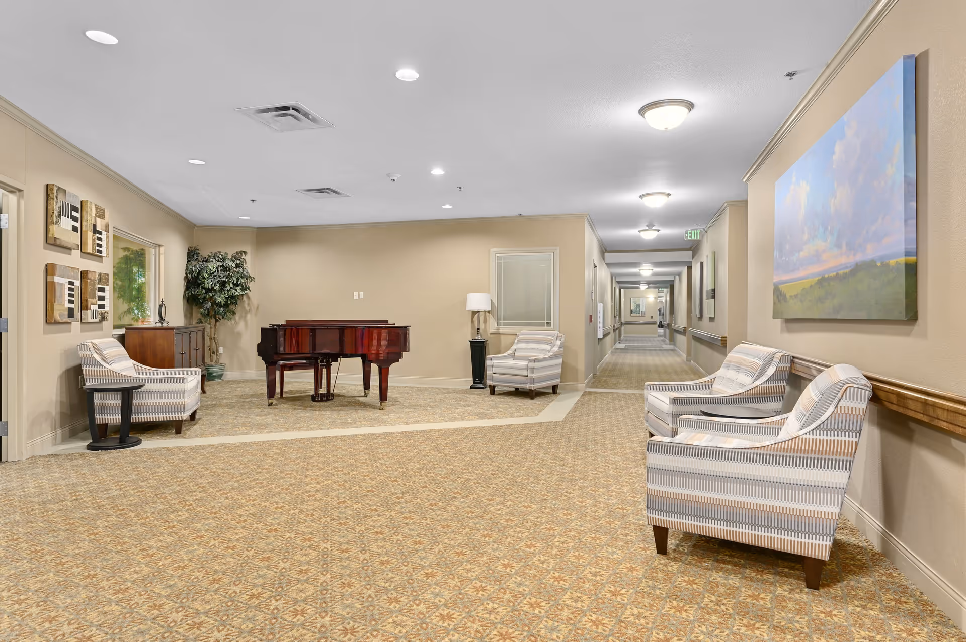 A spacious hallway area in a senior living facility with patterned carpet and beige walls. The area features a grand piano, several striped upholstered armchairs, a small side table, a floor lamp, and framed artwork on the walls. The hallway extends into the distance with multiple ceiling lights and handrails along the walls.