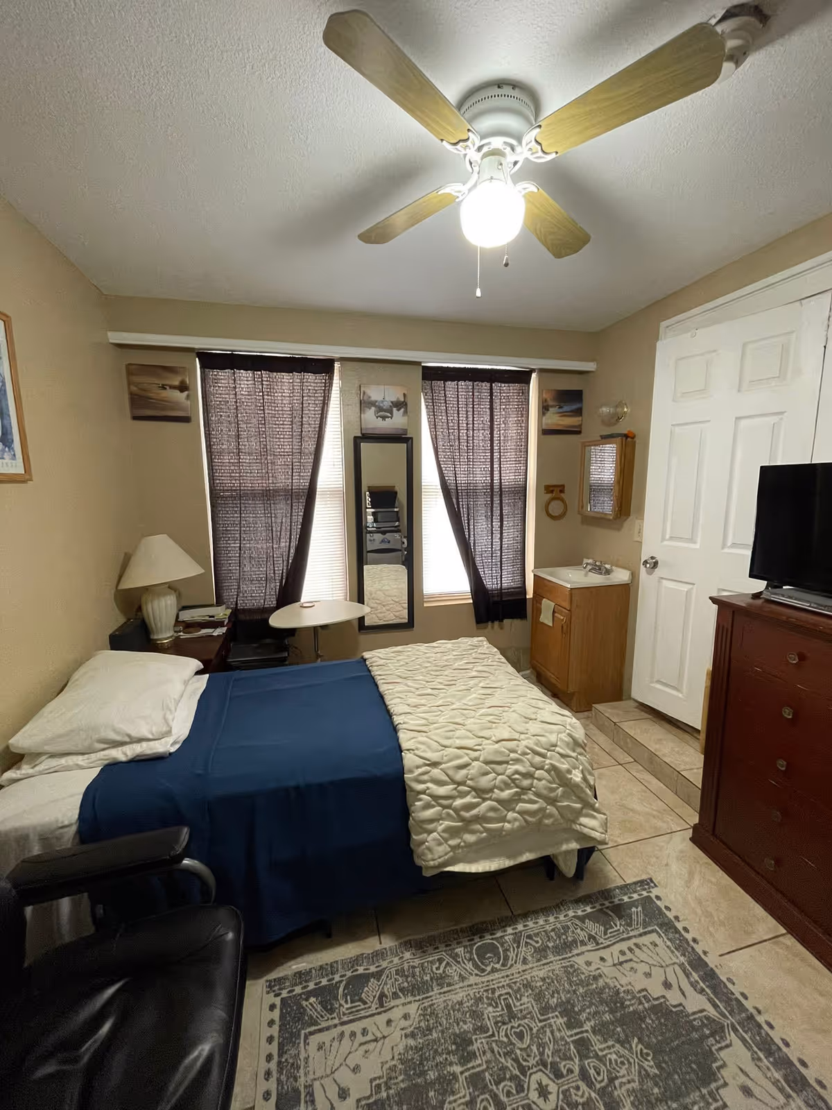 A small bedroom with a single bed covered with a blue sheet and a white quilt. There is a black leather chair in the foreground, a wooden dresser with a TV on top, and a small sink with a mirror above it near a white door. The room has beige walls, a ceiling fan with a light, and two windows with dark curtains. A patterned rug covers part of the tiled floor.