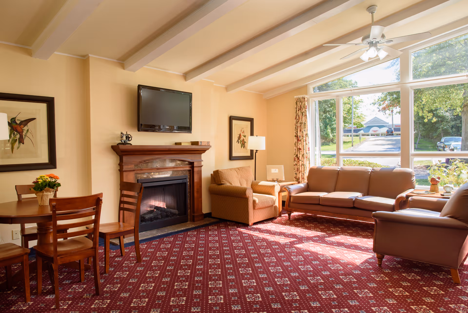 Sunlit living room with sofas, an armchair, a fireplace with a TV mounted above, a small dining table, and large windows looking outside.