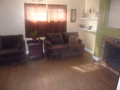 Cozy living room with brown sofa and chair, plant, shelves, and brick fireplace.