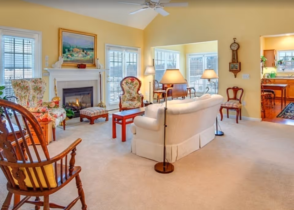A cozy living room with beige carpet and yellow walls, featuring a white sofa, floral upholstered armchairs, a wooden rocking chair, a fireplace with a painting above it, several lamps, and large windows letting in natural light. The room opens into a kitchen area with wooden cabinets and a dining table.