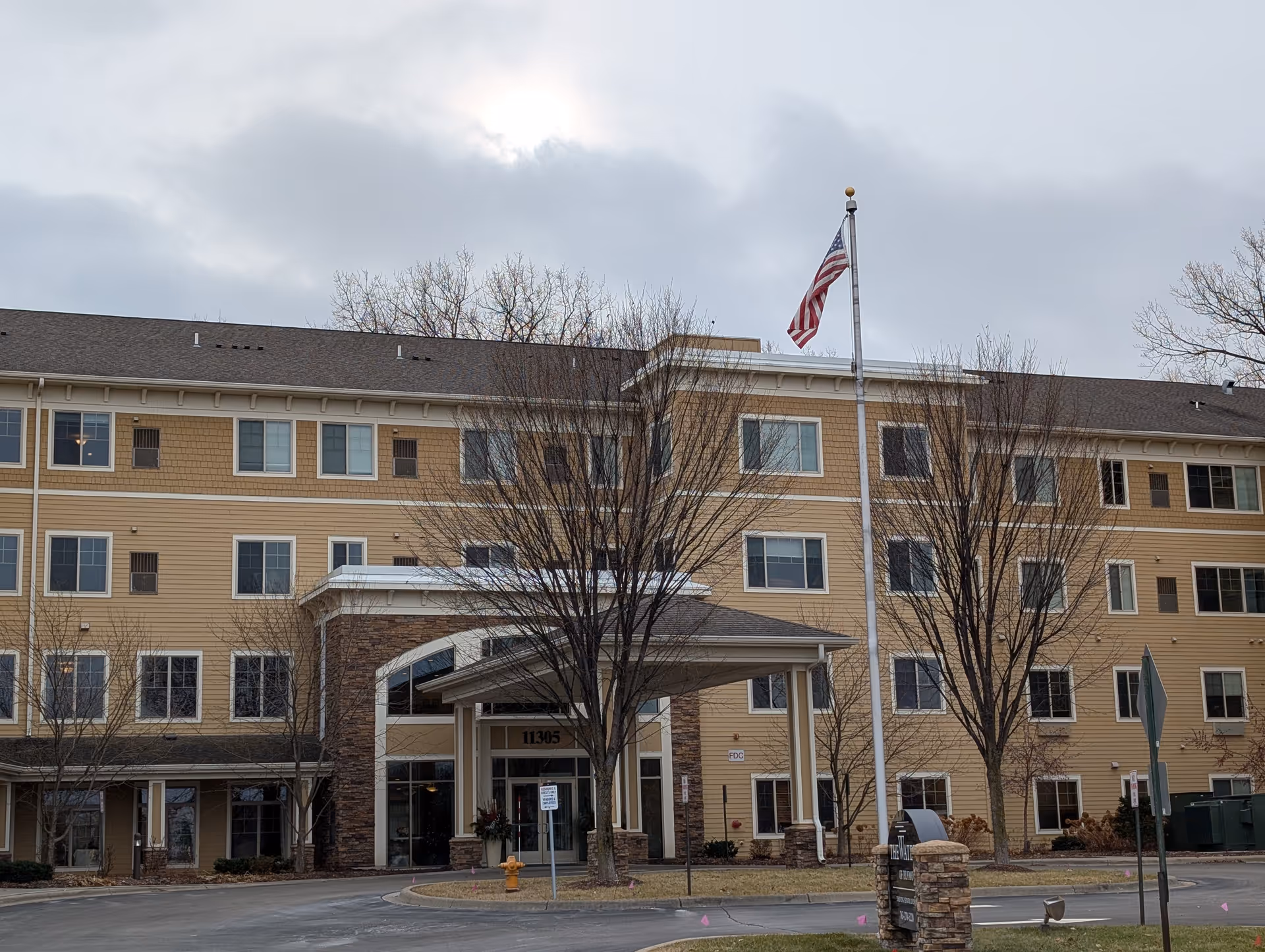 Exterior view of a multi-story senior living facility building with beige siding and stone accents. The entrance has a covered drop-off area with the number 11305 above the door. There are leafless trees in front of the building and an American flag flying on a flagpole near the entrance. The sky is cloudy.