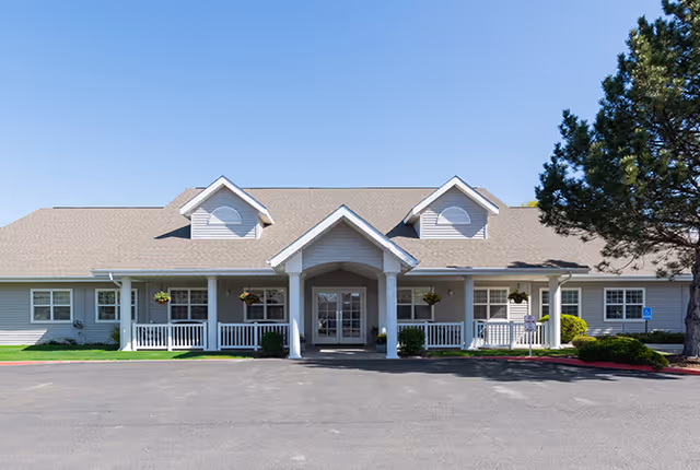 Front exterior view of a single-story senior living facility building with a covered entrance, white railings, hanging flower baskets, and a large tree on the right side under a clear blue sky.