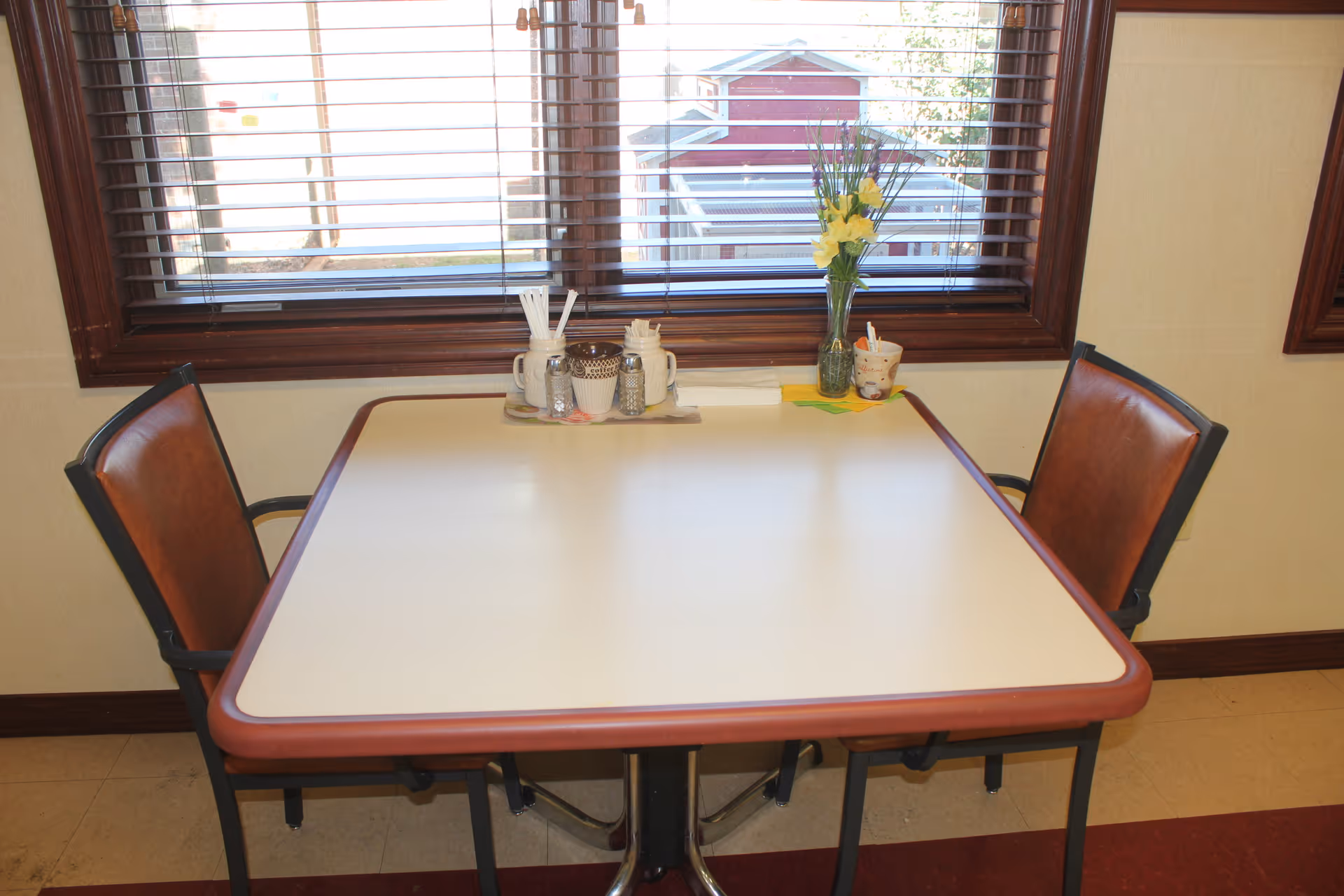 A small dining table with two chairs positioned on either side. On the table, there is a tray holding condiments, napkins, and utensils, along with a vase containing yellow flowers. Behind the table is a window with wooden blinds, showing a partial view of a red building outside.