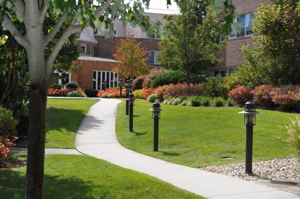 A winding concrete pathway through a well-maintained garden area with green grass, trees, and shrubs leading to a brick building with multiple windows.