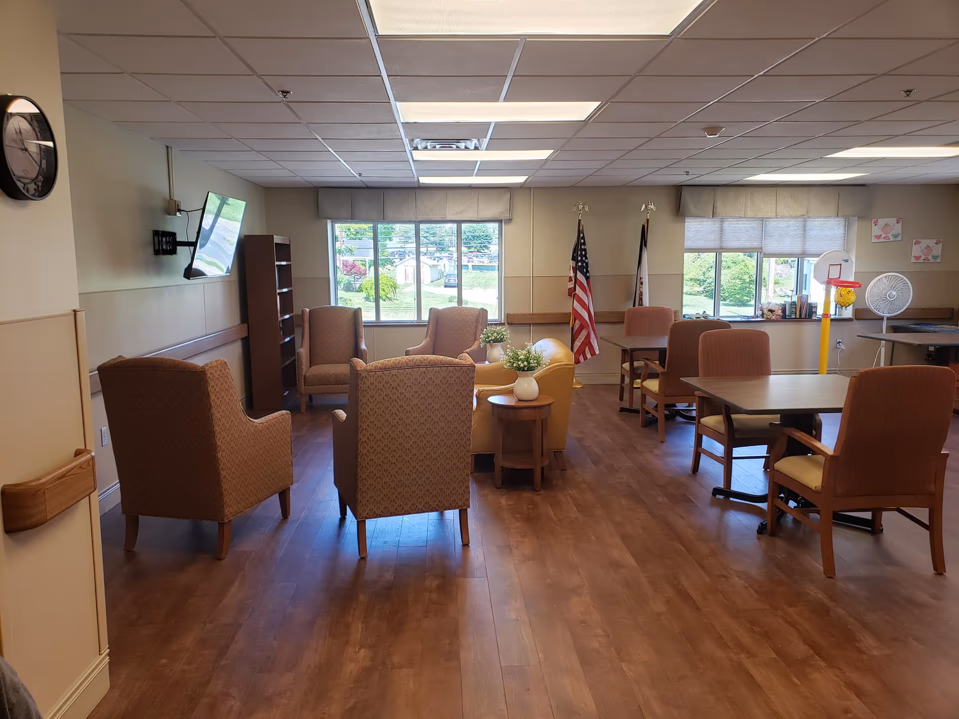A spacious common area in a senior living facility with several cushioned armchairs arranged around small tables with potted plants. There are larger tables with chairs on the right side, a wall-mounted TV on the left, and two flags near the windows at the back. The room has large windows letting in natural light and wood-style flooring.