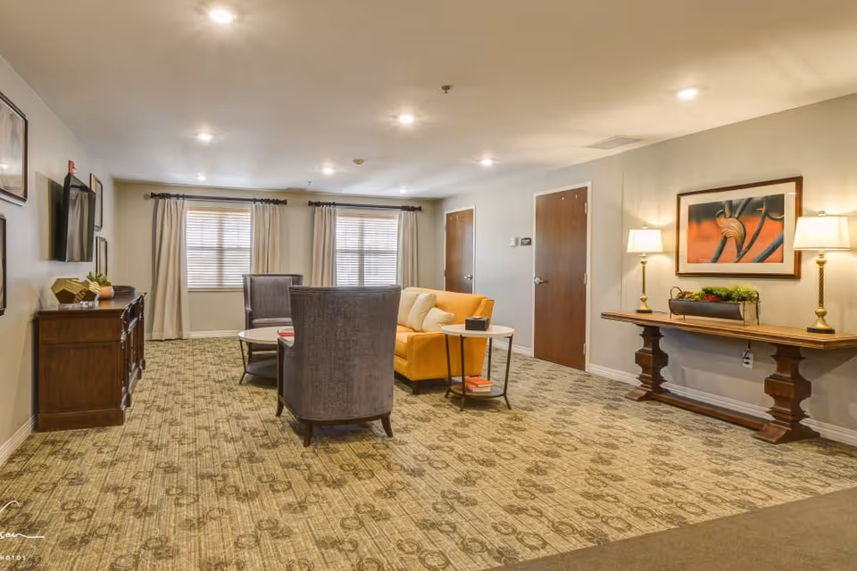 A well-lit living room area in a senior living facility featuring a yellow sofa with white pillows, two gray armchairs, a round coffee table, and a side table with books. The room has patterned carpet, beige walls, two windows with curtains, a wooden console table with two lamps and a decorative plant, and framed artwork on the walls.