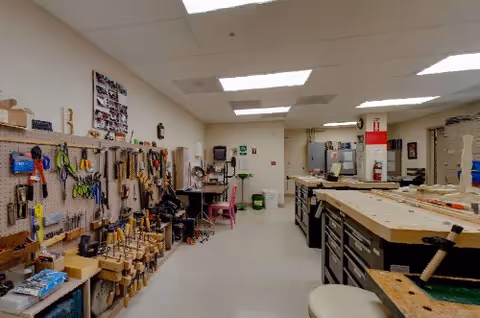 Interior view of a well-organized workshop room with various woodworking tools hanging on pegboards along the left wall and workbenches with drawers on the right side. The room is brightly lit with ceiling lights and has a clean, spacious floor.