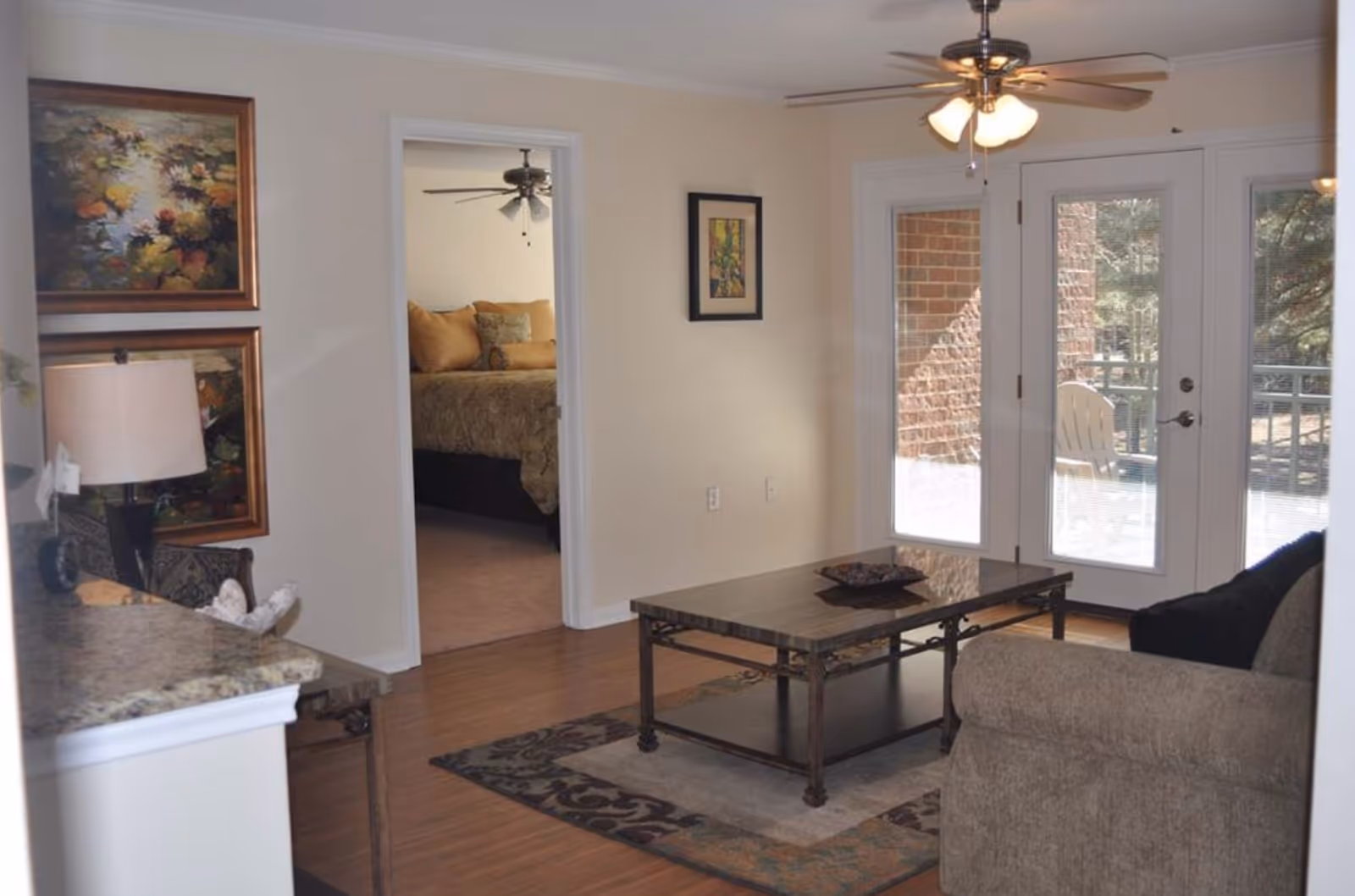 A cozy living room area with a beige couch, a decorative rug, and a rectangular coffee table. The room has wooden flooring and a ceiling fan with lights. There are glass doors leading to an outdoor patio with a chair visible outside. A doorway leads to a bedroom with a bed and ceiling fan. The walls are decorated with framed artwork and a table lamp is placed on a granite countertop.