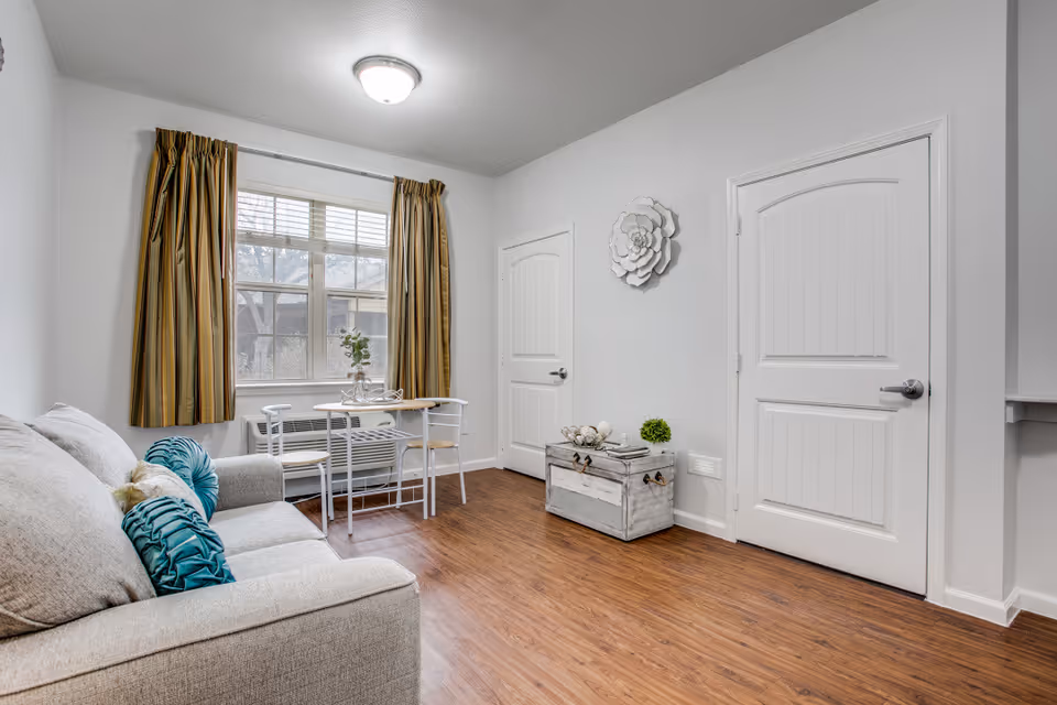 Bright living room with a sofa and teal pillows, a small table and chairs by a window, hardwood floors, wall decor, and two white doors.