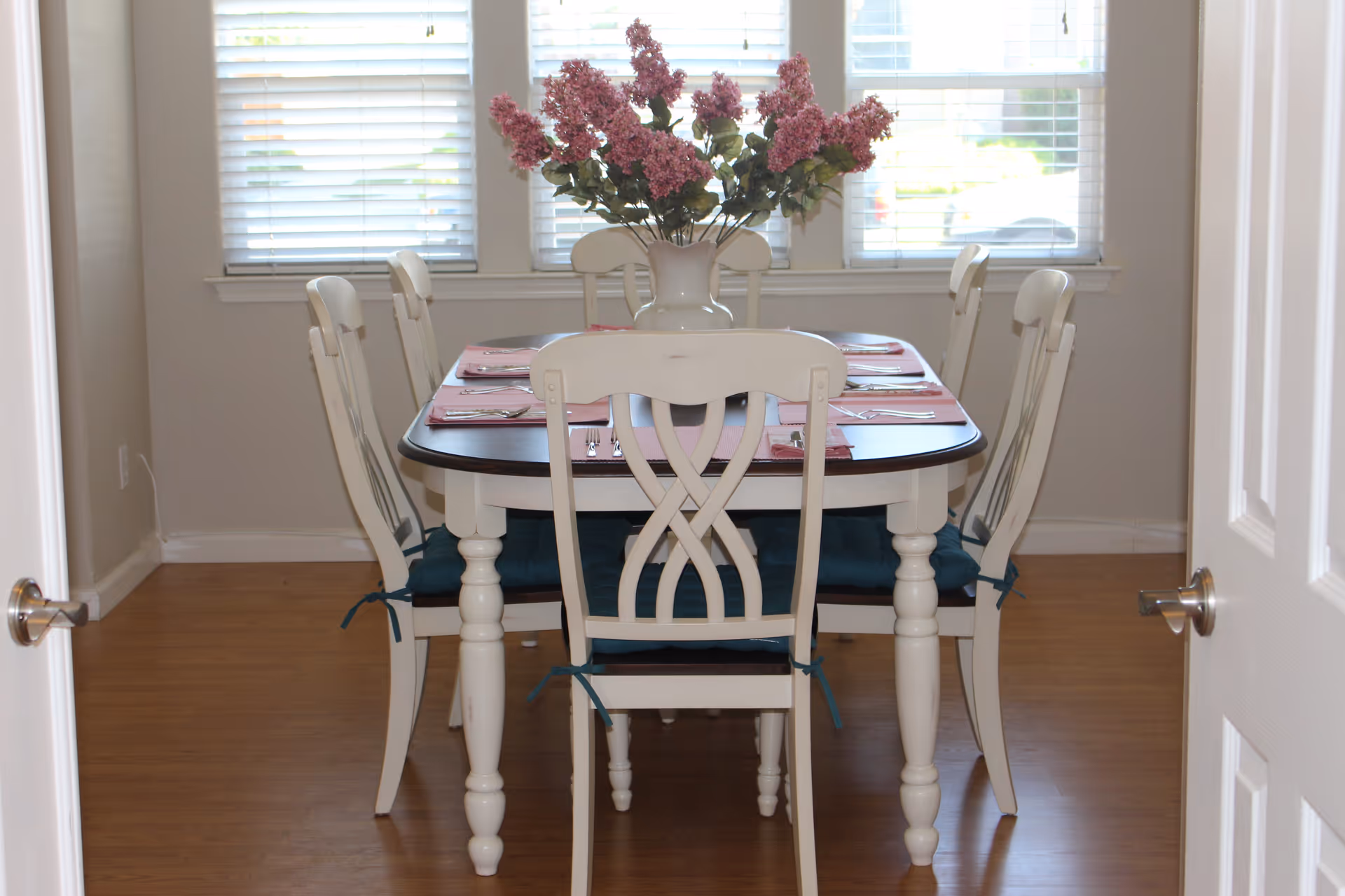 A dining room with a wooden table set for six people, featuring white chairs with blue cushions. A vase with pink flowers is placed in the center of the table. The room has wooden flooring and three windows with white blinds in the background.