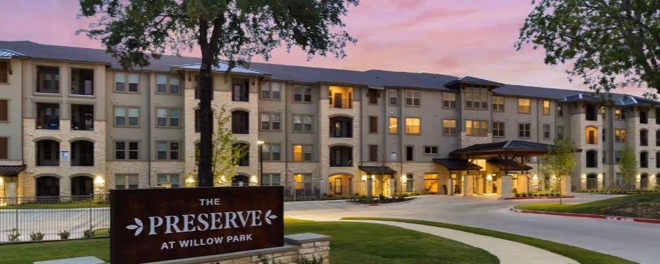 Front exterior of The Preserve at Willow Park senior living building with its illuminated entrance and sign in the foreground at dusk.