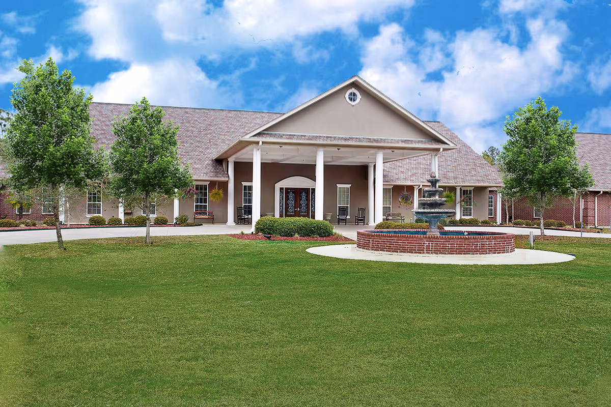 Front exterior view of a senior living facility building with a covered entrance supported by white columns, a circular brick fountain in front, green lawn, and several trees under a partly cloudy blue sky.