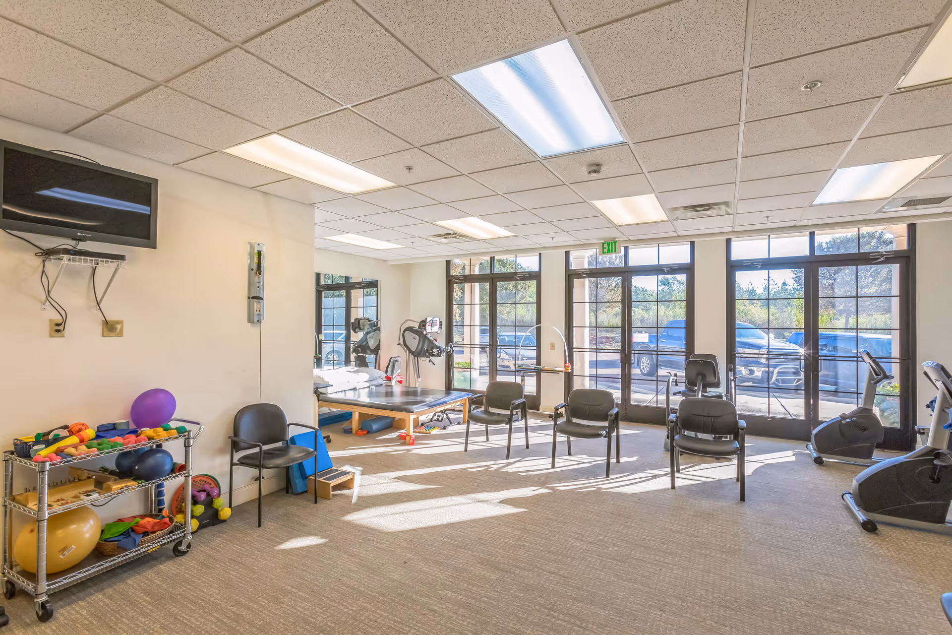 A bright exercise room with large windows letting in natural light. The room contains exercise equipment including stationary bikes, a therapy table, chairs arranged in a row, and a rack with various colorful exercise balls and weights. A wall-mounted TV is visible on the left side.