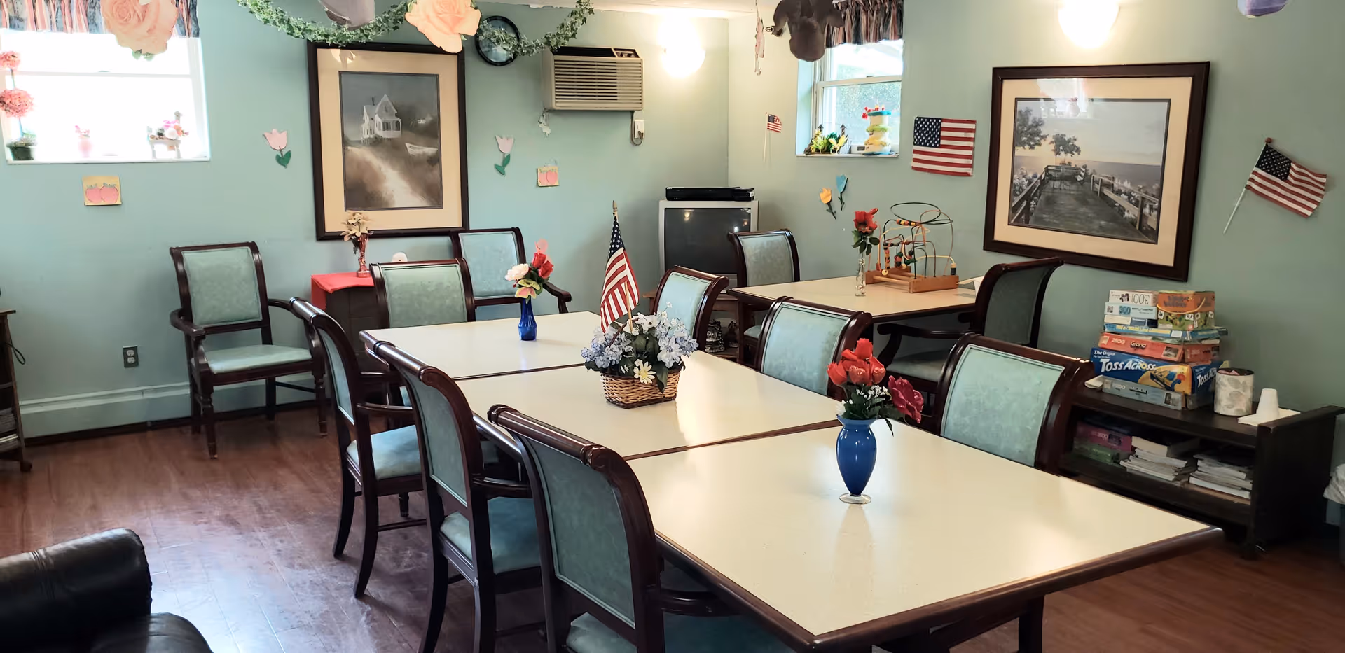 A well-lit dining room with several tables arranged together, surrounded by chairs with green upholstery. The tables have small flower vases and an American flag centerpiece. The walls are light green with framed pictures, small American flags, and decorative flowers. There is a window air conditioning unit, a small TV, and a shelf with board games and books.
