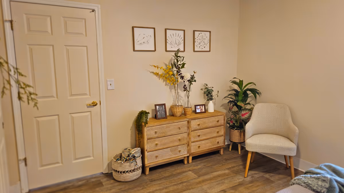 A cozy bedroom corner with a wooden dresser topped with plants and photos, framed art above, and an upholstered chair.