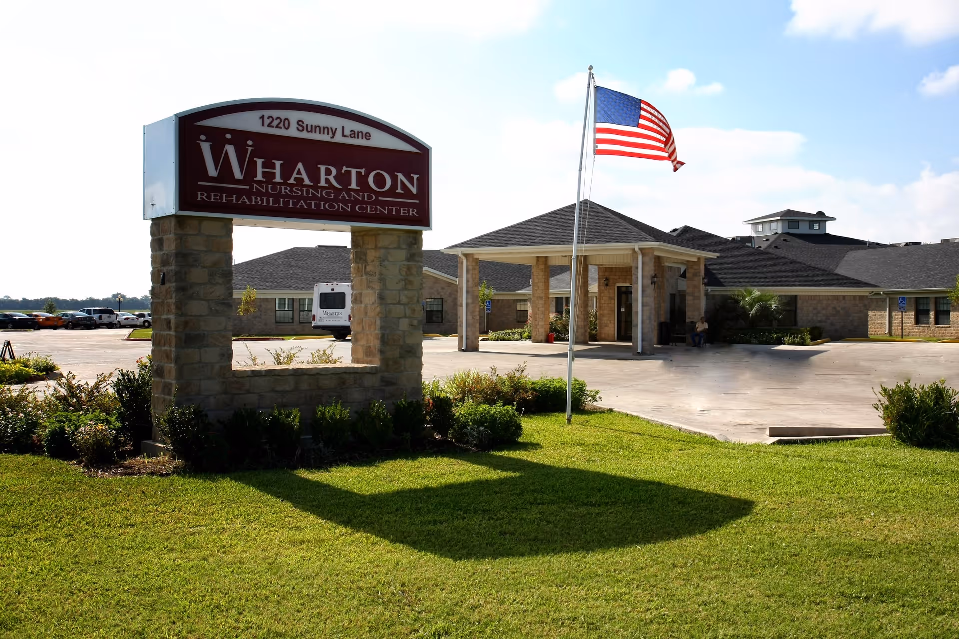 Sign reading 'Wharton Nursing and Rehabilitation Center' by the lawn with an American flag and the facility entrance in the background.