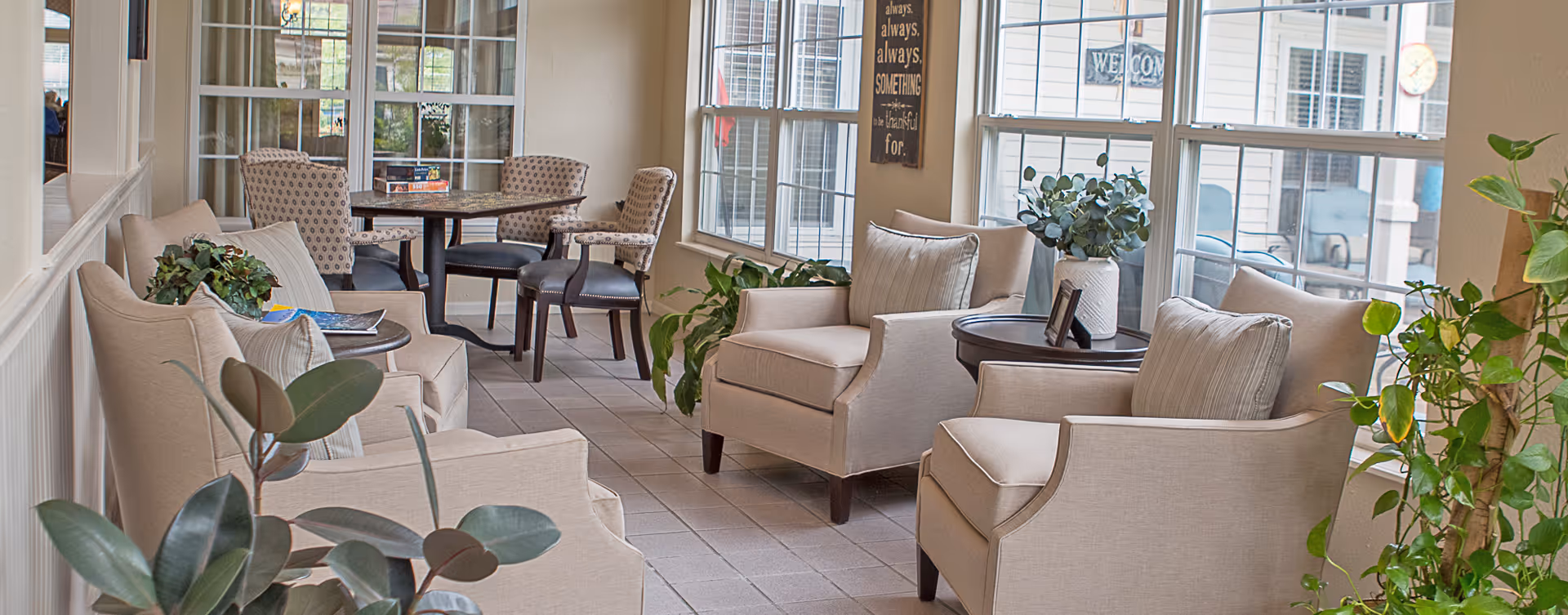 A bright and cozy sitting area with beige armchairs and small round tables, surrounded by large windows letting in natural light. There are several green plants placed around the space, and a dining table with chairs is visible in the background.