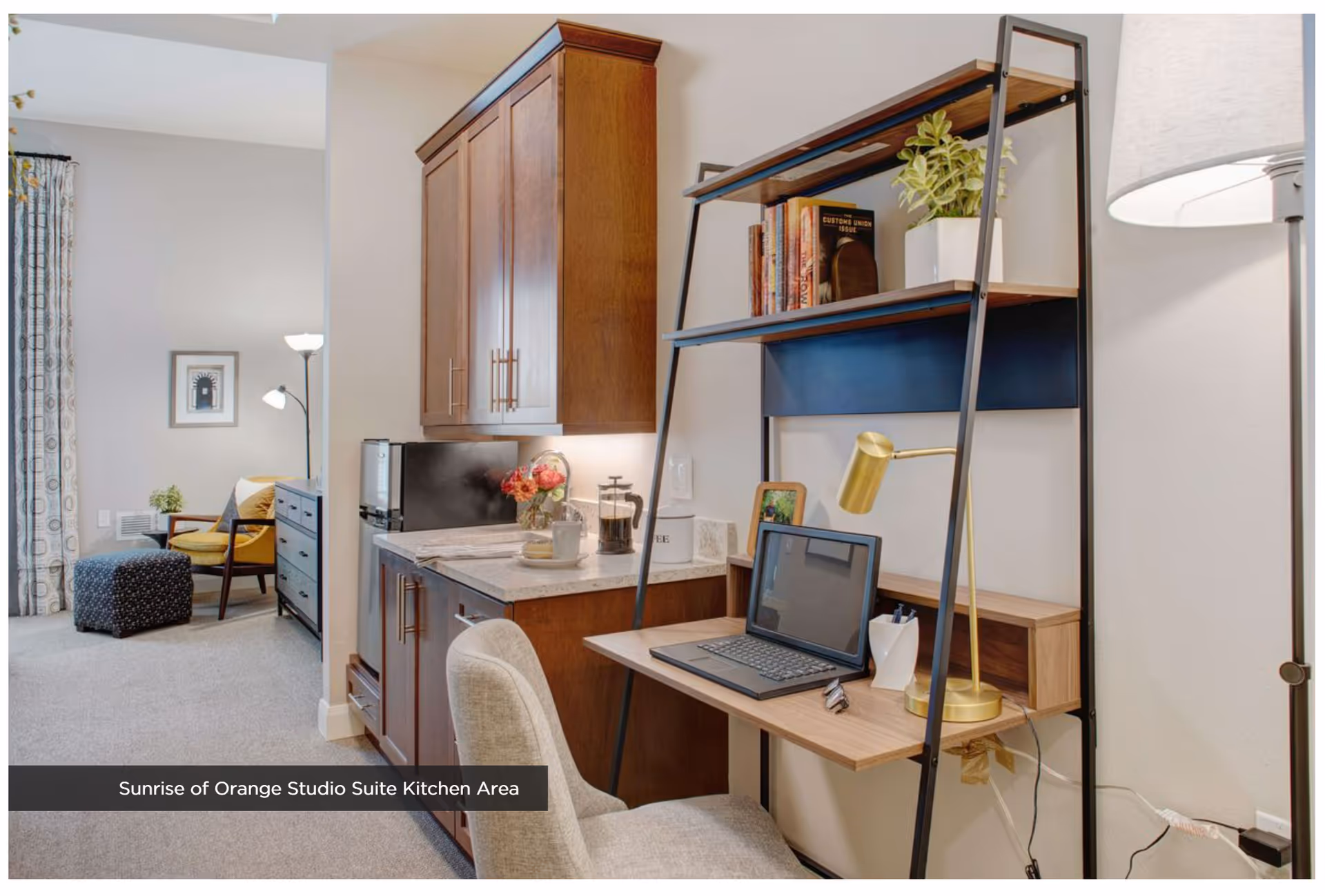 A studio suite kitchen area with wooden cabinets, a countertop with a coffee maker and flowers, and a small desk with a laptop, gold desk lamp, and decorative items. In the background, there is a cozy sitting area with a yellow chair, a patterned ottoman, a floor lamp, and a dresser with a plant on top.