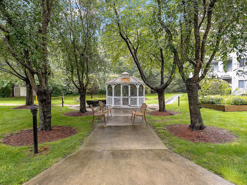A peaceful outdoor garden area with a concrete pathway leading to a white gazebo surrounded by trees and green grass. There are several metal chairs placed along the pathway, and a multi-story building is partially visible on the right side.