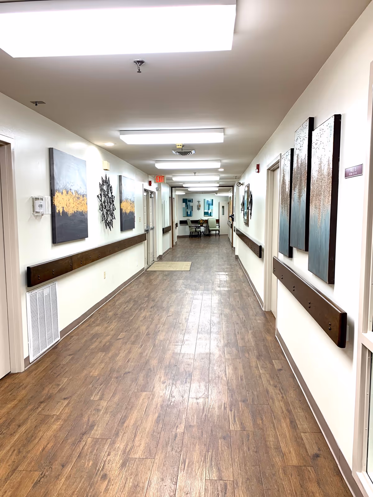 A long, well-lit hallway with wood-patterned flooring and white walls. The walls are decorated with abstract art pieces and metal wall sculptures. Handrails run along both sides of the hallway. At the end of the hallway, there is a small seating area with chairs and a table. Ceiling lights illuminate the corridor evenly.