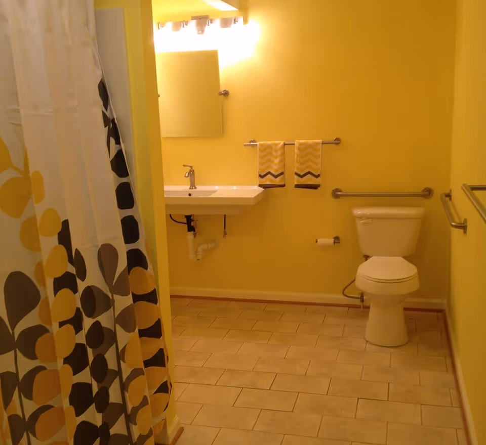 A bathroom with yellow walls and beige tiled floor. It features a white toilet with grab bars on both sides, a wall-mounted sink with exposed plumbing, a mirror above the sink, and two yellow patterned hand towels hanging on a towel rack. A shower curtain with a yellow, black, and gray leaf pattern is partially visible on the left side.