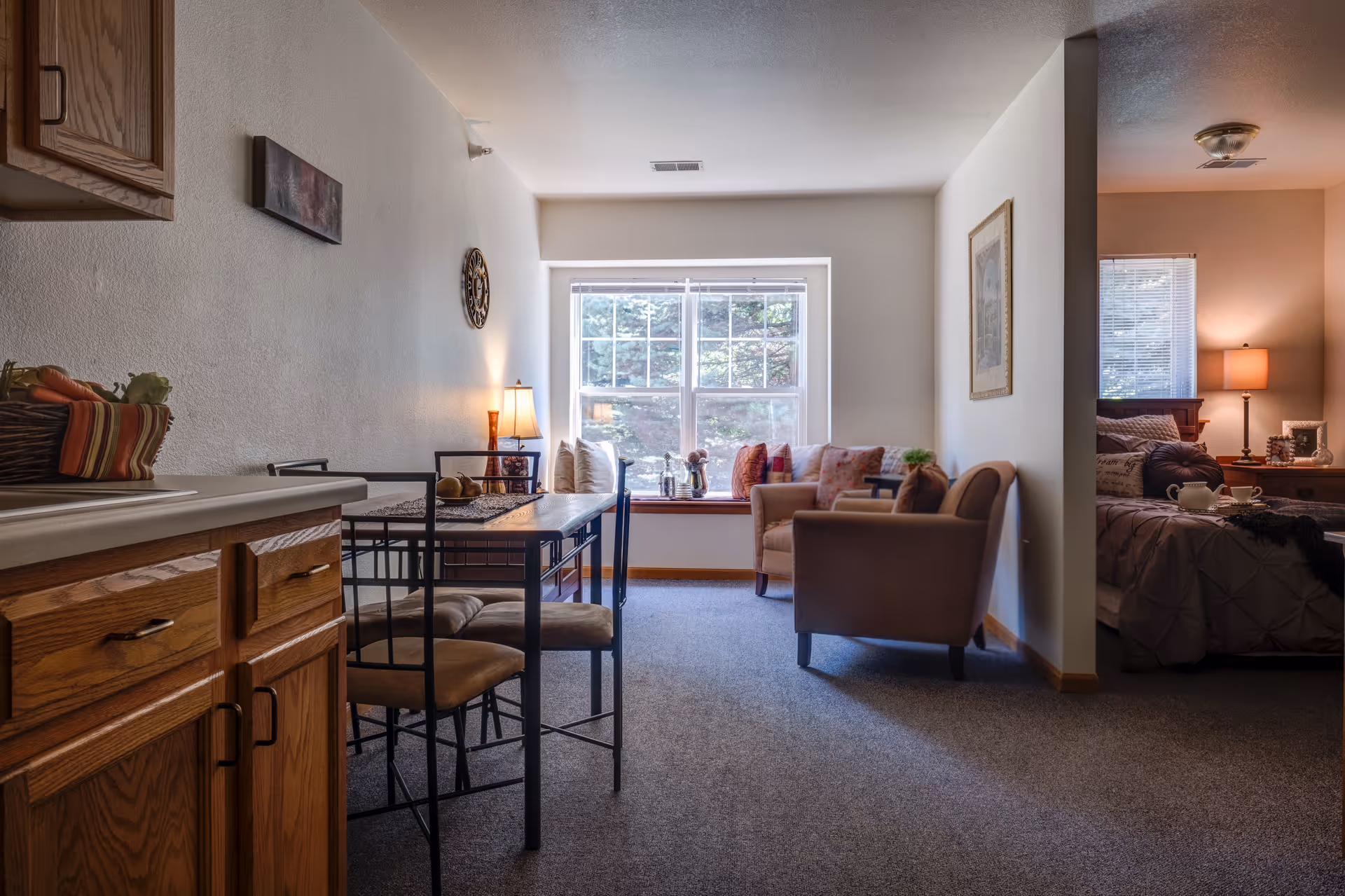 Interior view of a senior living facility apartment showing a small dining area with a table and chairs next to a kitchen counter with wooden cabinets. Beyond the dining area is a cozy living space with armchairs, decorative pillows, a lamp, and a large window letting in natural light. To the right, a partially visible bedroom with a bed, nightstand, lamp, and window with blinds.