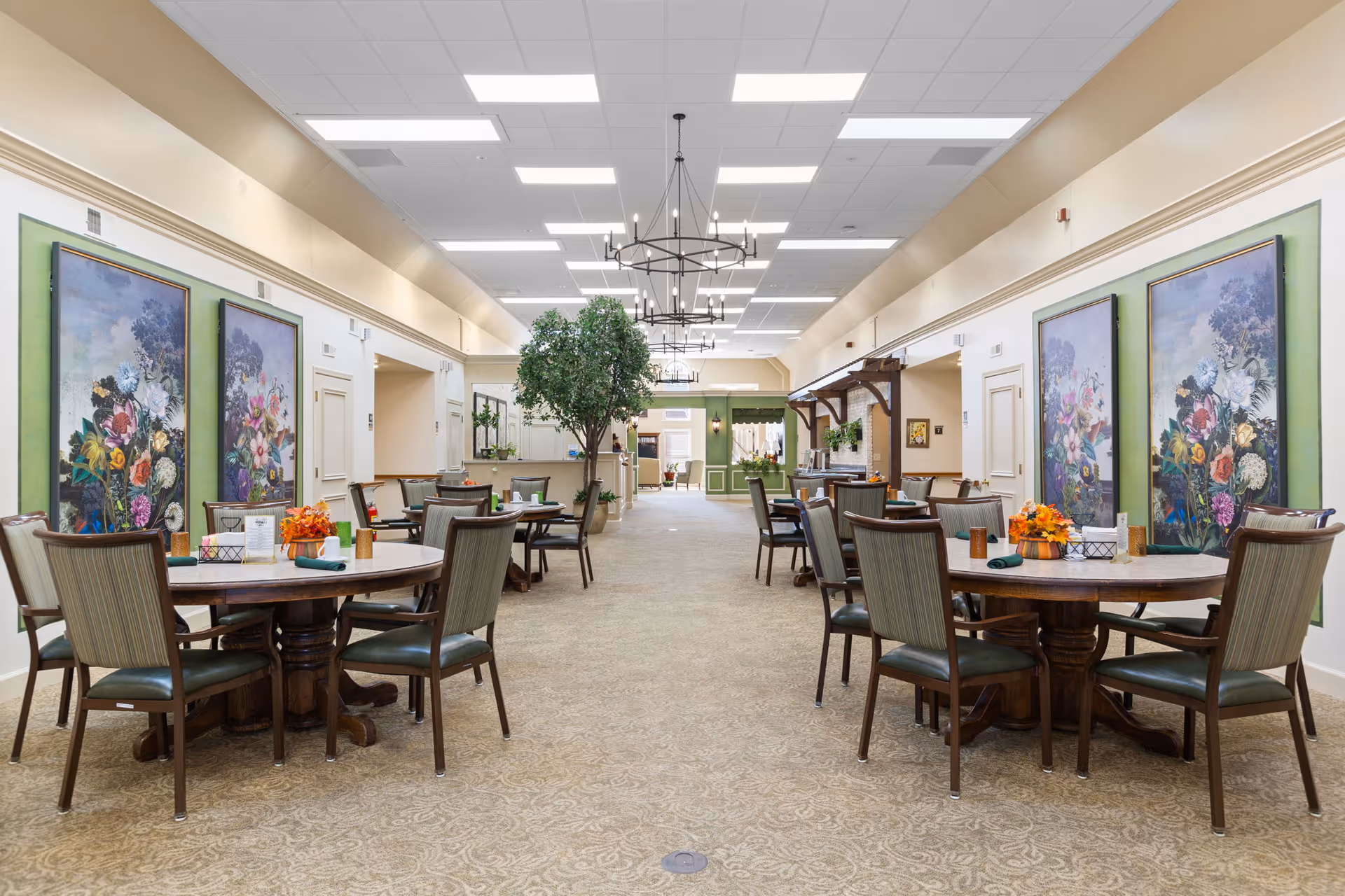 Spacious senior dining room with round tables and chairs, floral artwork on the walls, and a central walkway leading to a service area.