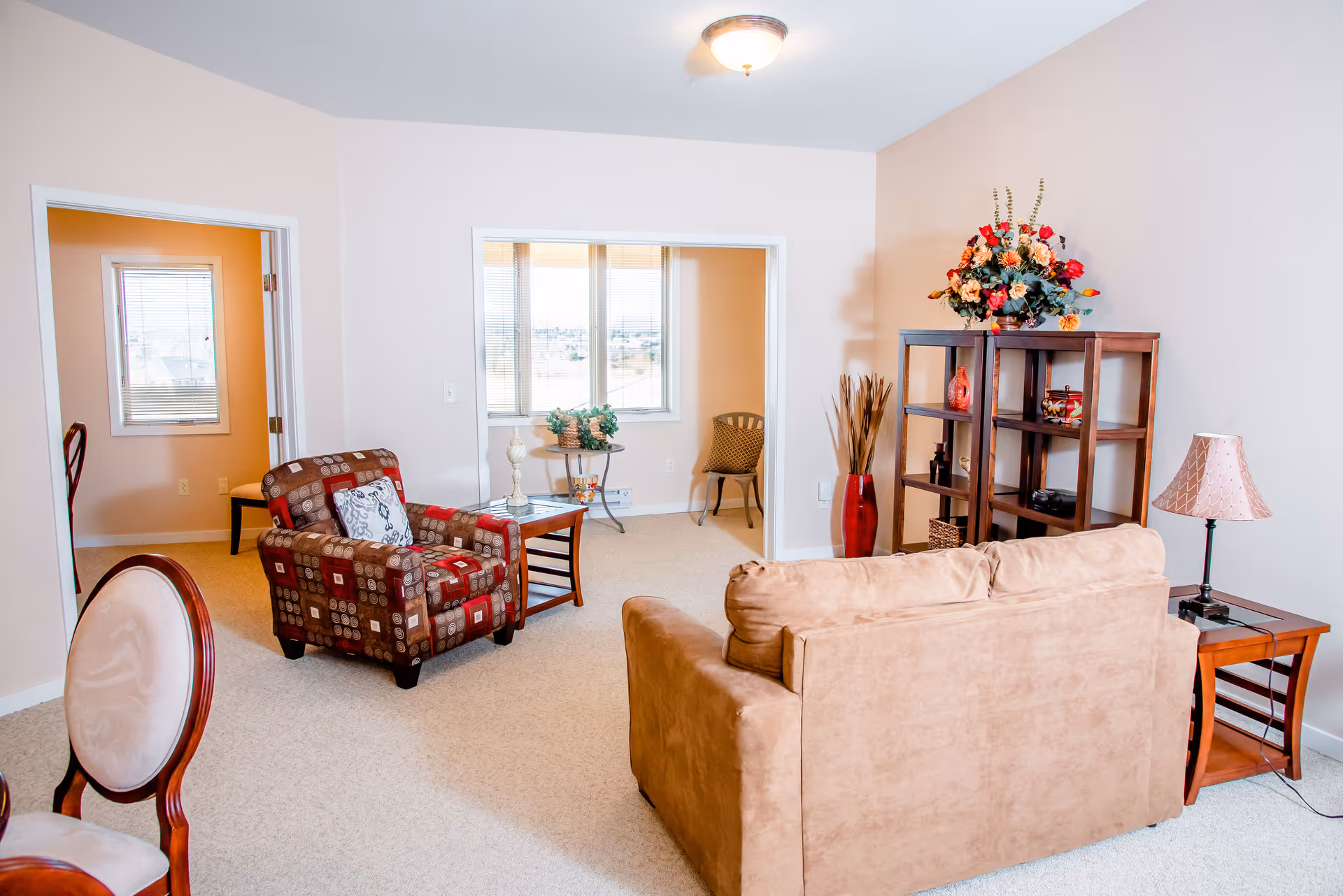 A bright and cozy living room in a senior living facility featuring a beige sofa, a patterned armchair with a decorative pillow, a wooden side table with a lamp, and a wooden shelving unit adorned with a large floral arrangement and decorative items. The room has light-colored walls and carpeted floors, with an adjacent room visible through an open doorway.