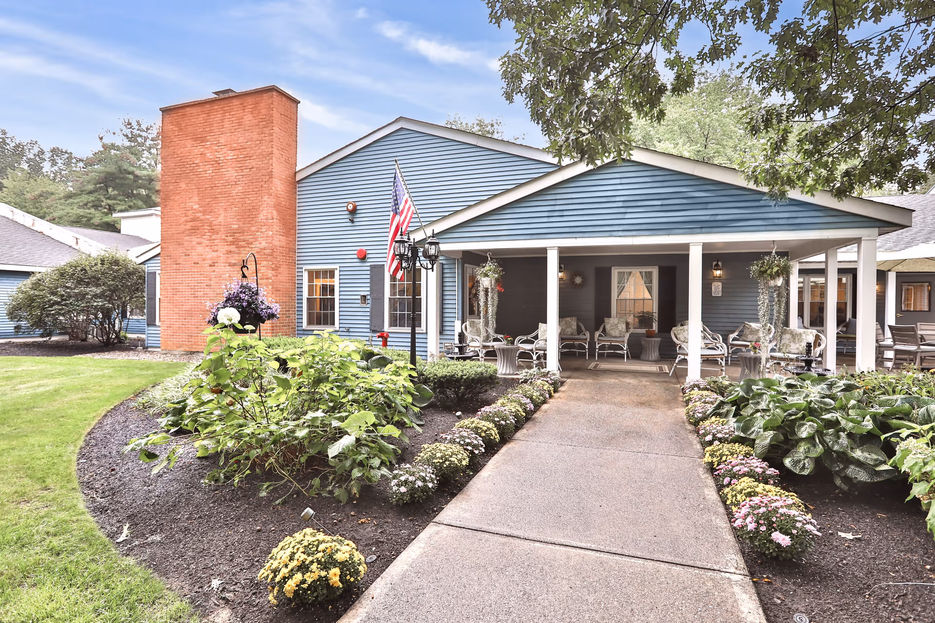 Exterior view of a senior living facility with a blue building featuring a covered porch with white columns. The porch has several chairs and hanging plants. There is a concrete walkway leading to the porch, bordered by flower beds with various plants and flowers. An American flag is displayed near the entrance, and a large brick chimney is visible on the left side of the building.