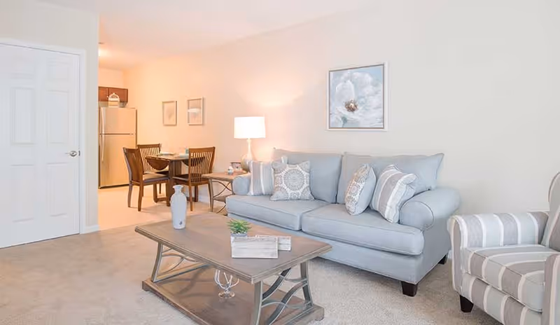 A bright and cozy living room with a light gray sofa adorned with patterned pillows, a striped armchair, and a wooden coffee table with decorative items. In the background, there is a small dining area with a table and four chairs, a lamp on a side table, and a kitchen area with a refrigerator and cabinets.
