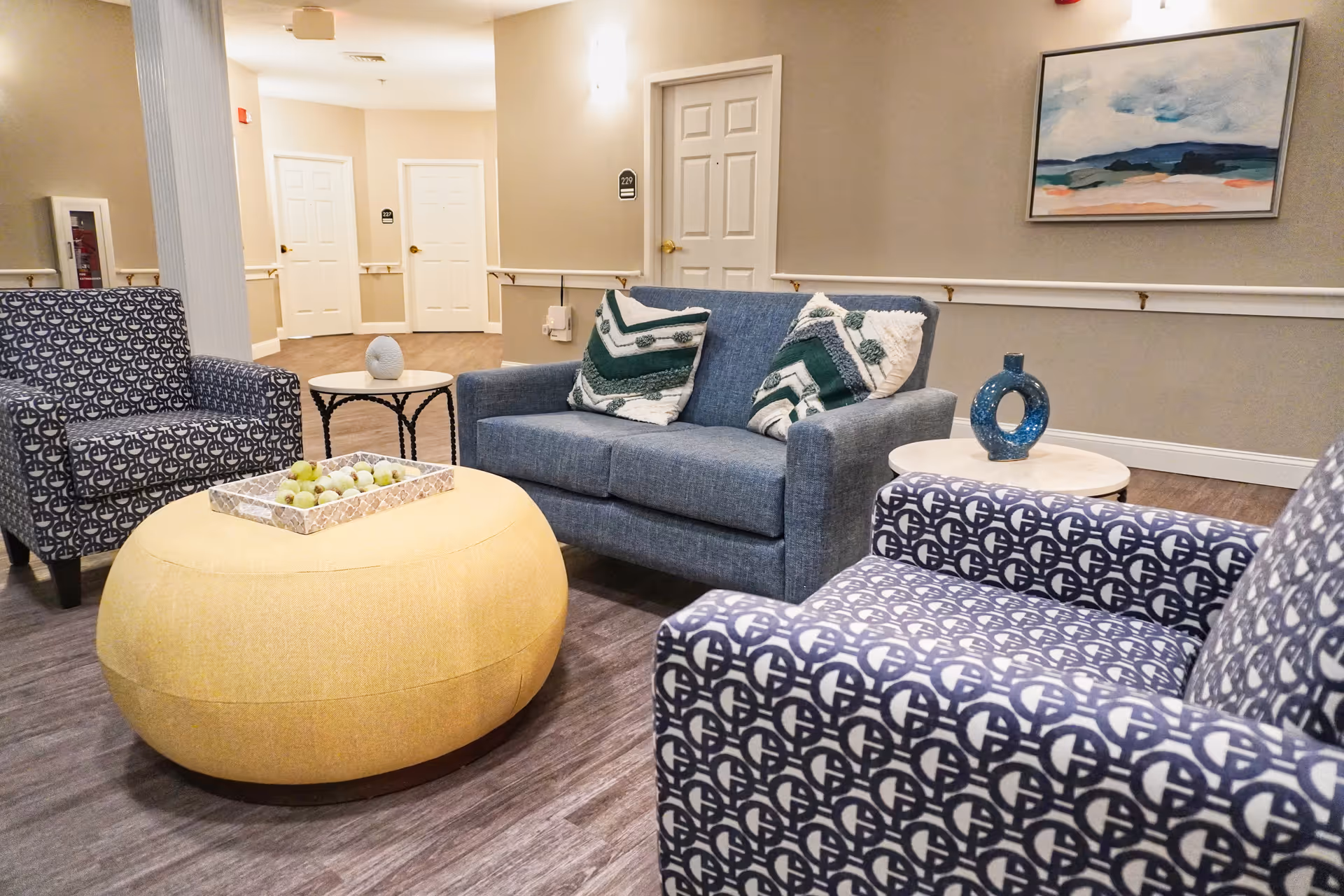 A cozy seating area in a senior living facility with a blue loveseat adorned with two green and white patterned pillows, two patterned armchairs, a large yellow ottoman with a tray of decorative green balls, and two small round side tables with decorative items. The background shows beige walls, white doors with room numbers, a framed abstract painting, and wooden flooring.