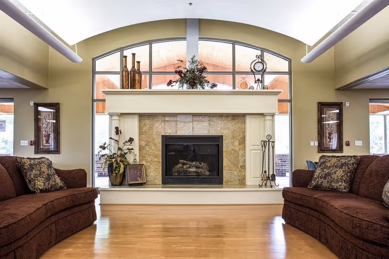A cozy living room area with two brown sofas facing a central fireplace. The fireplace has a stone surround and a white mantle decorated with vases, a clock, and floral arrangements. Large arched windows behind the fireplace allow natural light to fill the room. The walls are painted a soft beige color and the floor is wooden.
