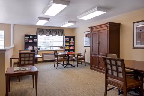 A well-lit common room with tables and chairs, bookshelves, and a large wooden cabinet near a window.