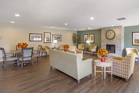 A spacious living room area in a senior living facility with multiple seating arrangements including sofas and armchairs around a fireplace with a large clock above it. There are several tables with flower vases, framed pictures on the walls, and a wooden floor.