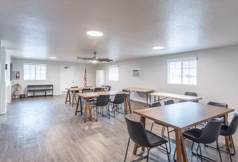 Bright multipurpose dining/activity room with several wooden tables and black chairs, windows, and an American flag.