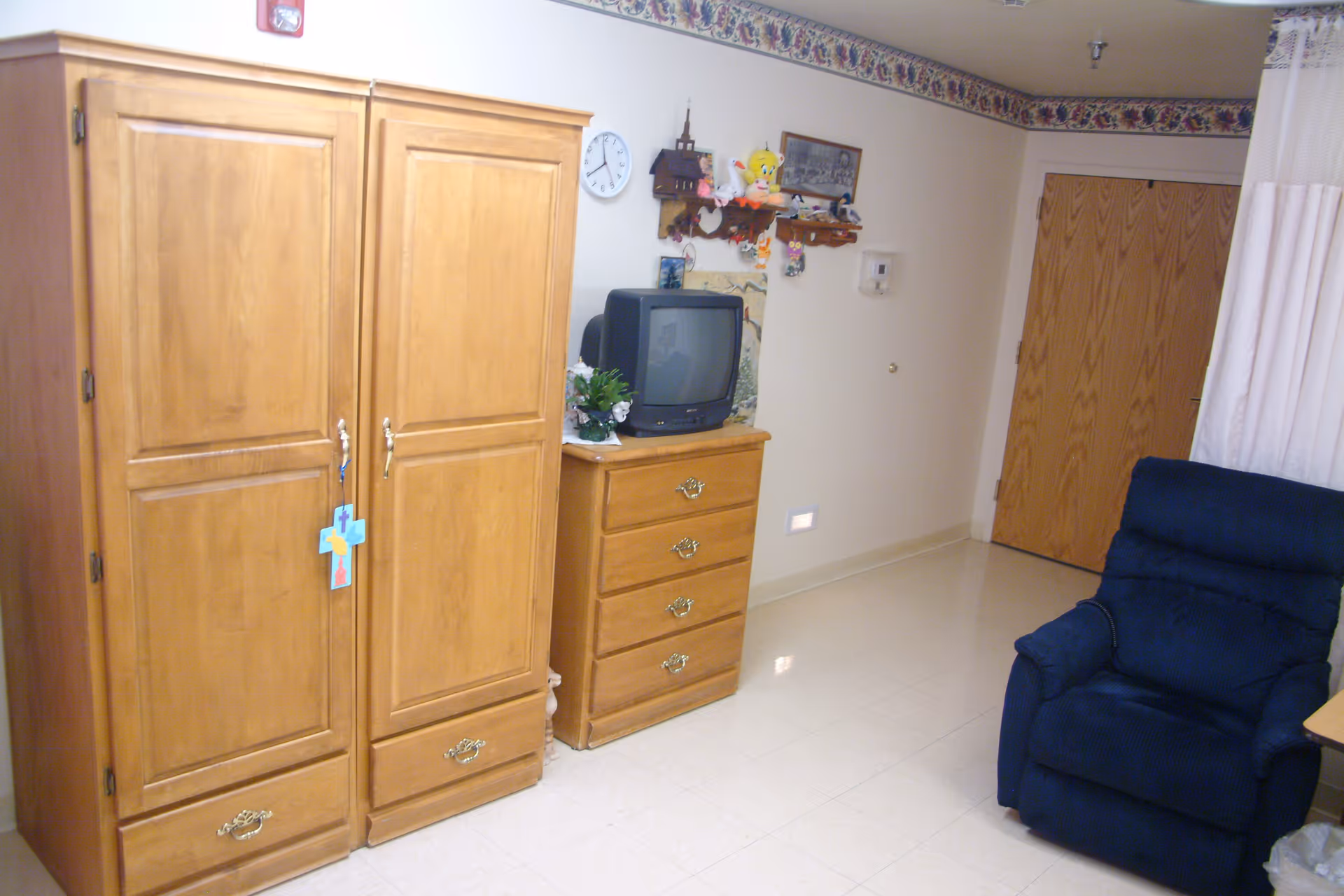 Small resident room interior with wooden armoire and dresser holding a TV, a blue recliner, and a closed door.