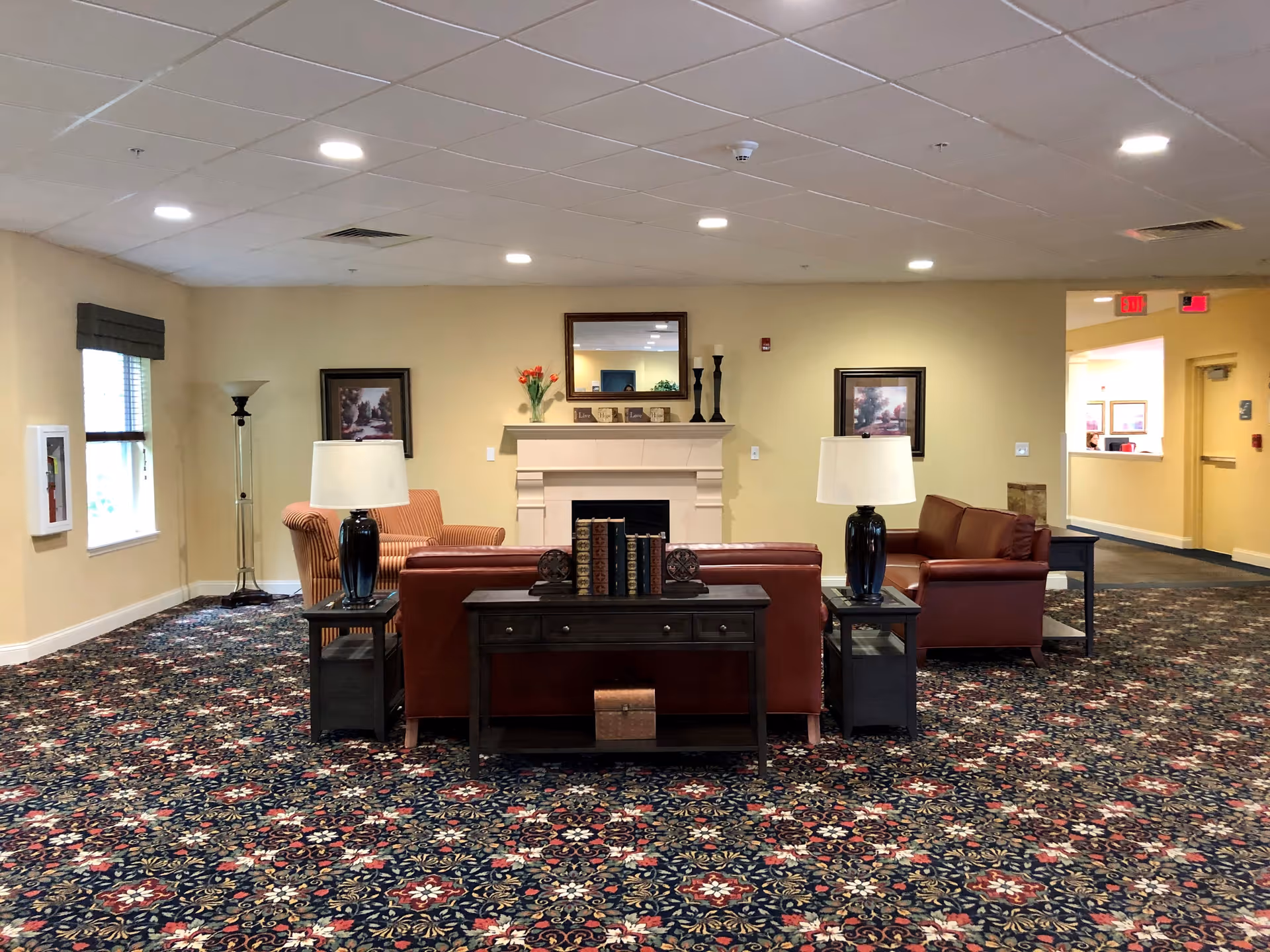 A cozy senior living facility common area with patterned carpet, two leather sofas, two side tables with lamps, a fireplace with a mirror and decorative items above it, and framed artwork on the walls. There is a window with a valance on the left and a hallway leading to other rooms on the right.