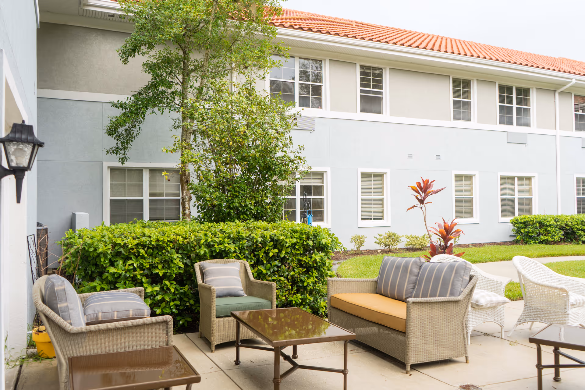 Outdoor patio area with cushioned wicker chairs and sofas arranged around glass-top tables, surrounded by green bushes and trees, adjacent to a two-story building with multiple windows and a red-tiled roof.