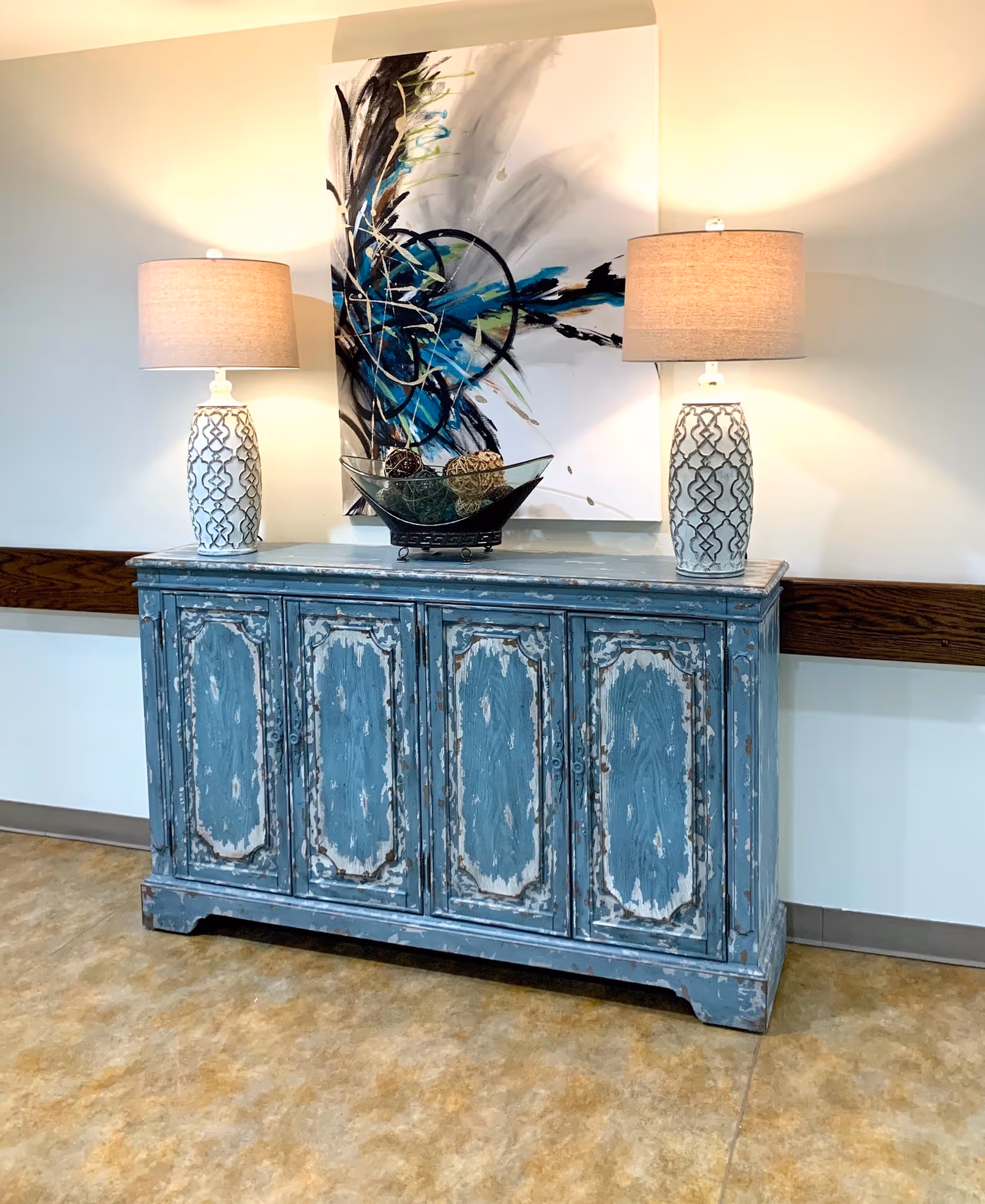 A distressed blue wooden cabinet with four doors stands against a white wall. On top of the cabinet are two matching table lamps with beige shades and white bases featuring a geometric pattern. Between the lamps is a decorative glass bowl containing three textured spheres. Above the cabinet hangs an abstract painting with black, blue, and white brushstrokes.