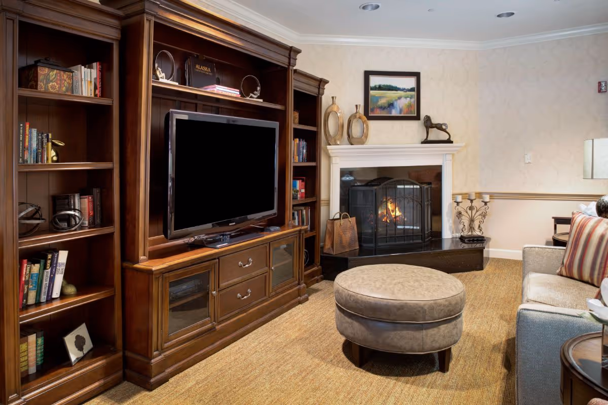 A cozy living room featuring a large wooden entertainment center with a flat-screen TV, bookshelves filled with books and decorative items, a lit fireplace with a black screen, a round beige ottoman, a beige sofa with striped cushions, and a side table with a lamp. The walls have a subtle patterned wallpaper and a framed landscape painting above the fireplace.