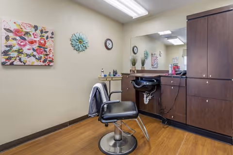 Interior view of a small salon or grooming area with a black salon chair in front of a large mirror. The room has wooden flooring and beige walls decorated with a colorful floral painting and a decorative wall clock. There is a countertop with hair care products and a cabinet for storage.