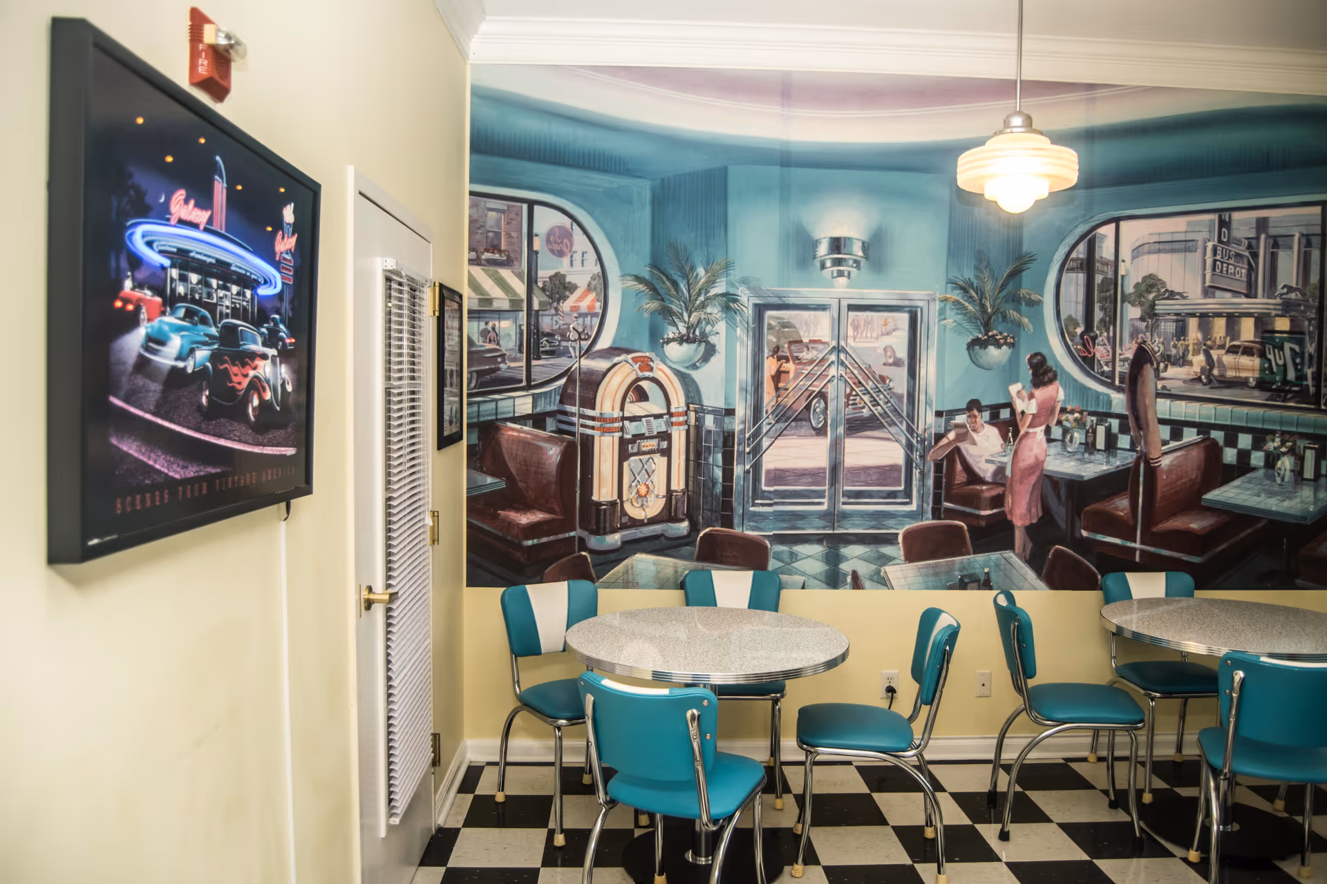 A retro-style dining area with turquoise and white chairs around round tables. The walls feature a large mural depicting a 1950s diner scene with a jukebox, people sitting in booths, and vintage cars outside. The floor has a black and white checkered pattern, and there is a framed neon-lit picture of classic cars on the adjacent wall.