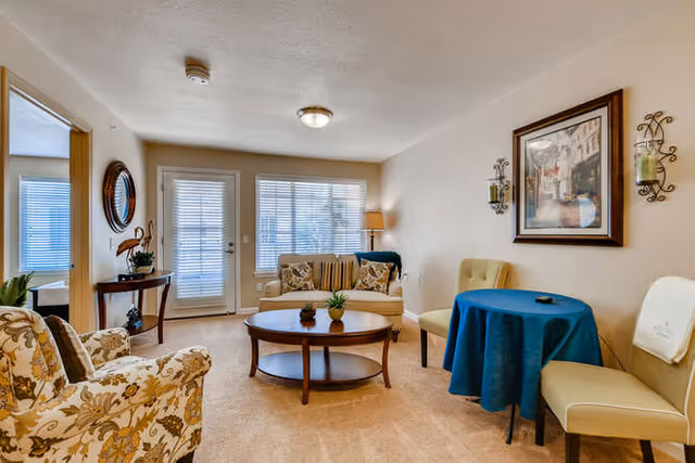 A cozy living room with beige walls and carpet. The room features a floral patterned armchair, a small round wooden coffee table with plants on it, a loveseat with striped and floral cushions, and a small round table covered with a blue tablecloth surrounded by two beige chairs. There is a framed painting on the wall above the table, two wall-mounted candle holders, a floor lamp near the loveseat, and a door and window with blinds letting in natural light.