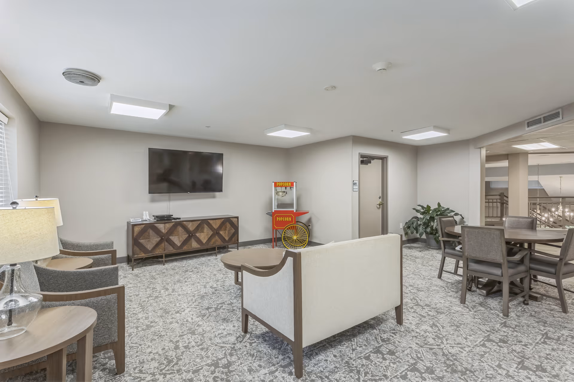 A common area in a senior living facility featuring a wall-mounted flat screen TV above a wooden console, a vintage-style popcorn machine, a beige loveseat, two gray armchairs with wooden frames, a round wooden coffee table, a dining table with six chairs, and a potted plant near a doorway. The room has patterned carpet flooring and ceiling lights.