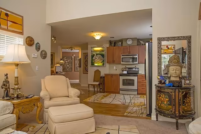 Interior view of a cozy living area with beige armchairs and ottoman, a wooden side table with a lamp and decorative items, and a kitchen in the background featuring wooden cabinets, a stove, microwave, and refrigerator. The room is decorated with framed artwork, a decorative cabinet with a Buddha head sculpture, and a large mirror.